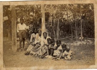 Heavily yellowed sepia photo of a family seated outdoors under banana trees
